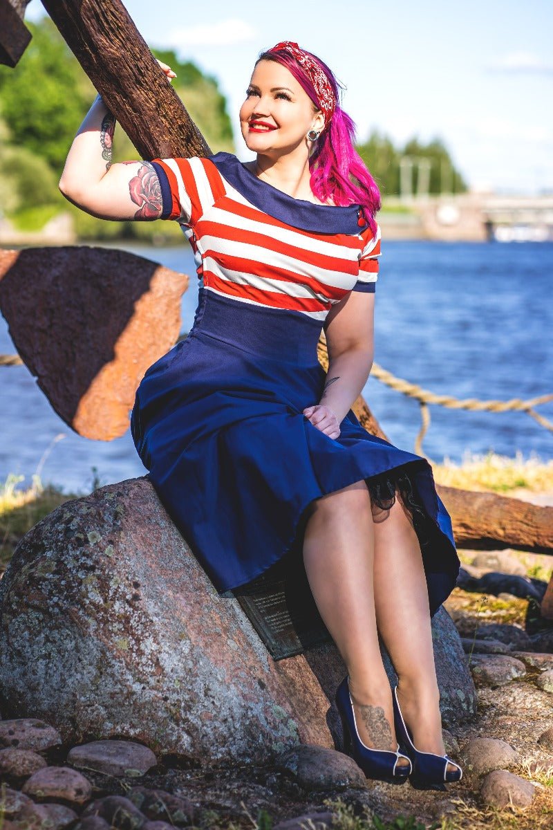 Woman wears our Darlene sailor swing dress in navy blue, with red and white stripes, in front of a boating lake