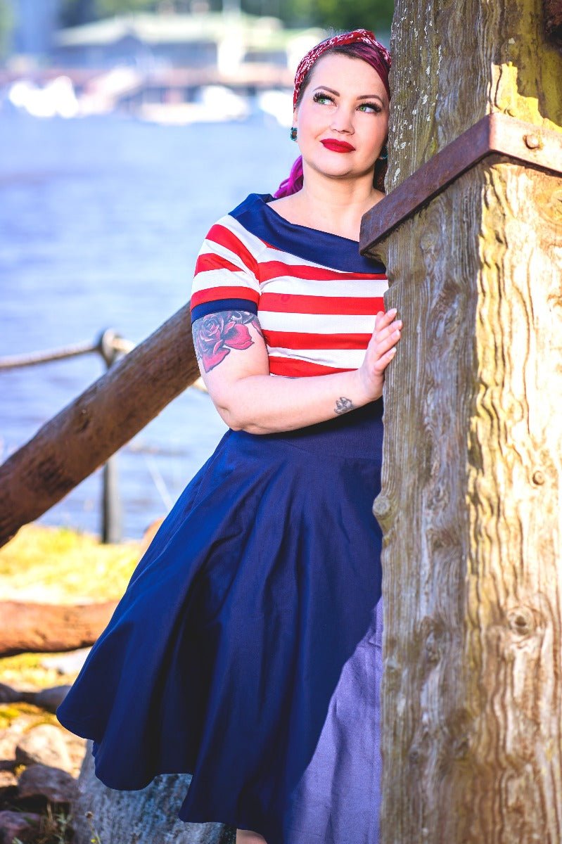 Woman wears our Darlene nautical dress in navy blue, with red and white stripes, in front of a boating lake