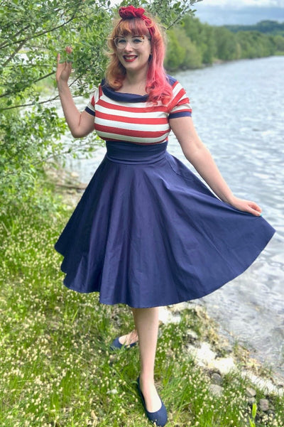 Woman wears our Darlene short sleeve dress in navy blue, with red and white stripes, in front of a lake