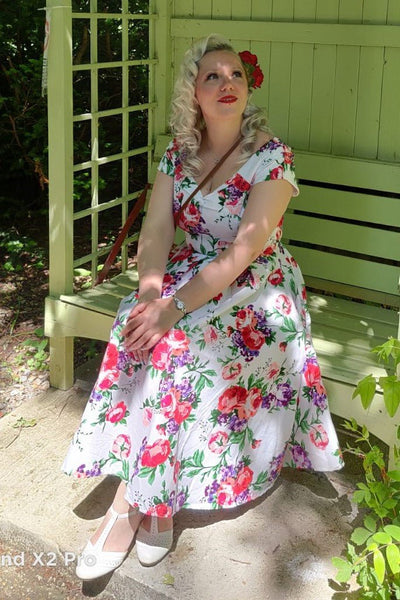 Woman wearing our Lily, off-shoulder 50's Evening Dress in White, with pink and purple flowers, sitting on a bench