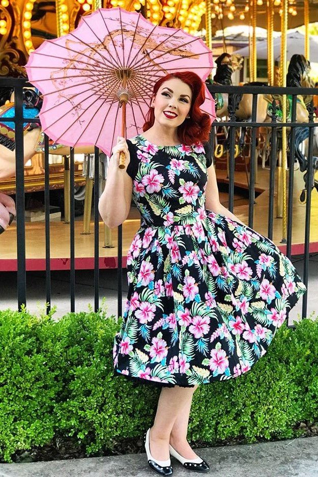 Woman wearing our Annie dress, in black/pink orchid floral print, with pink umbrella, in front of carousel
